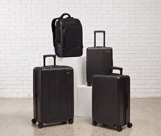 NOMATIC Bundles collection banner, a matching set of black hardside luggage and a backpack displayed against a white brick wall