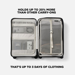 An open carry-on suitcase shown from above on a light gray background, with text above reading 'HOLDS UP TO 20% MORE THAN OTHER CARRY-ONS' and text below reading 'THAT'S UP TO 3 DAYS OF CLOTHING'. The suitcase has a black exterior, light gray interior lining, mesh organizer pockets, and a gray compression strap on the right compartment.