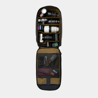 An open camera gear organizer bag shown from above on a light gray background. The bag is filled with camera lenses, lenses in a separate compartment, and various camera accessories and small items in the lower pocket.
