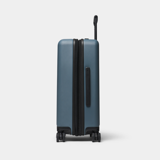 A hard-shell spinner suitcase in a muted slate blue color with black zippers, handles, and wheels, shown from a side profile on a light gray background.