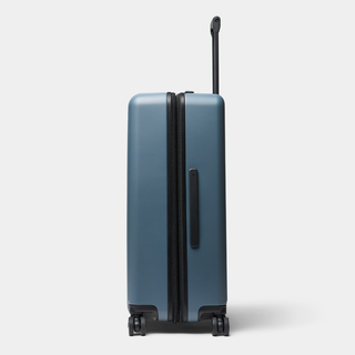 A hard-shell spinner suitcase in a muted slate blue color with black zippers, a vertical handle, and four wheels, shown on a light gray background.