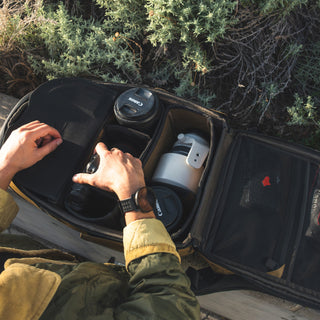 Person organizing coffee cups in a car's open storage compartment with greenery in the background.